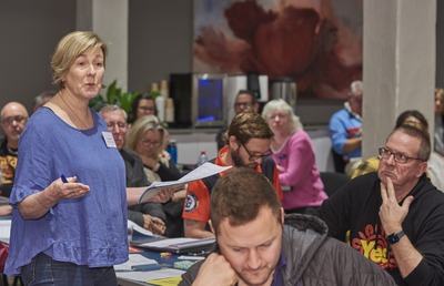 Standing woman speaks at Federation House during the TAFE review meeting while other participants watch and take notes.